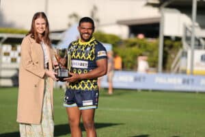 On the left is a woman in a brown coat, who is passing the Black & White Cabs Cup trophy to a man in a rugby jersey on the right. They are standing on the field.